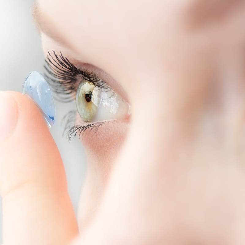 Gros plan sur une personne insérant une lentille de contact bleue dans un œil vert clair avec de longs cils. Scène de soin oculaire.
