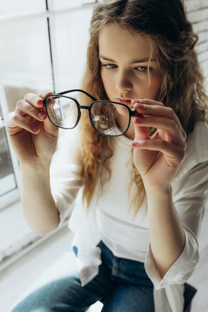 Jeune femme aux cheveux bouclés, vêtue d'une chemise blanche, examine attentivement des lunettes noires qu'elle tient entre ses mains. Des reflets sont visibles sur les verres.