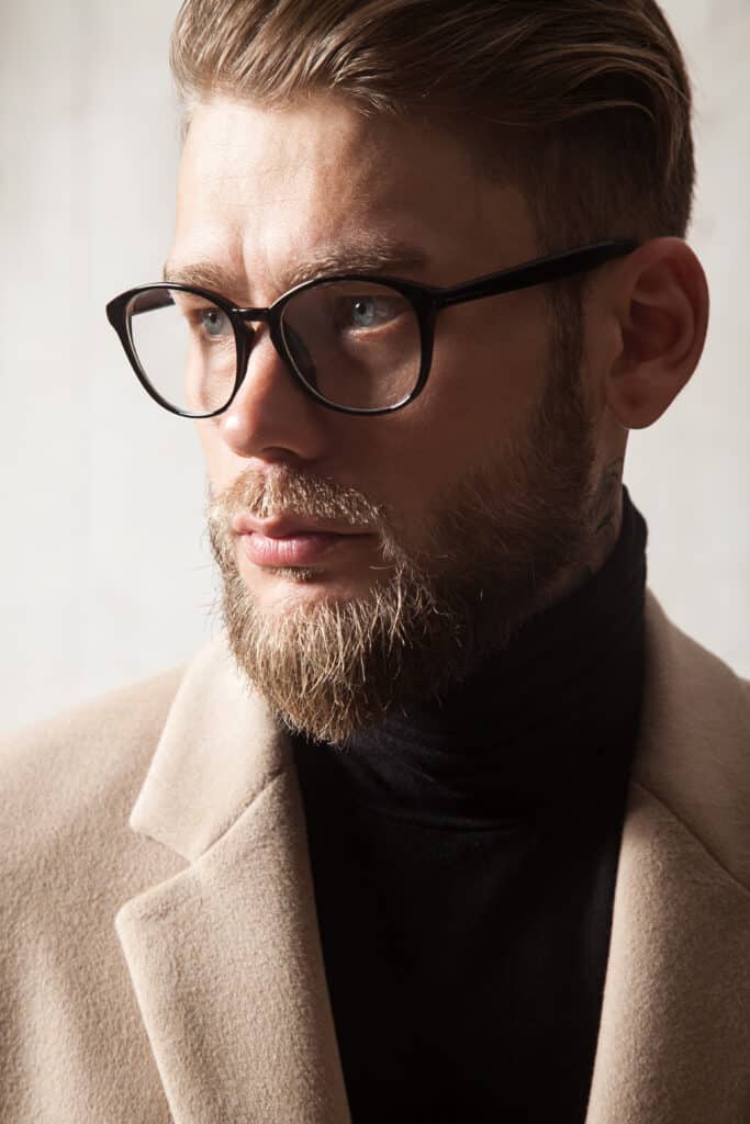 Portrait élégant d'un homme blond barbu aux yeux bleus, portant des lunettes noires et un manteau beige. Son regard est pensif.