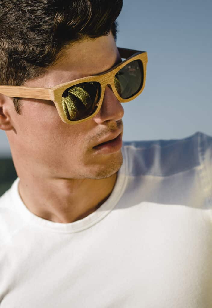 Jeune homme au profil distinct, portant des lunettes de soleil en bambou aux verres dorés miroirs, contemplant le paysage.