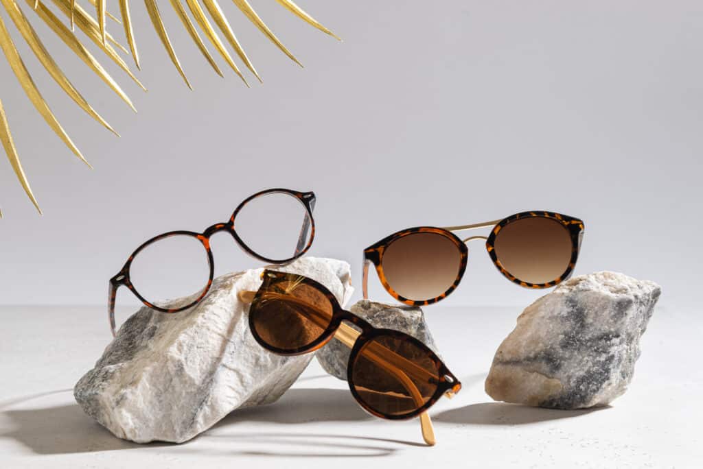 Trois paires de lunettes (vue et soleil) à monture écaille, posées sur des roches blanches, avec des feuilles de palmier dorées.