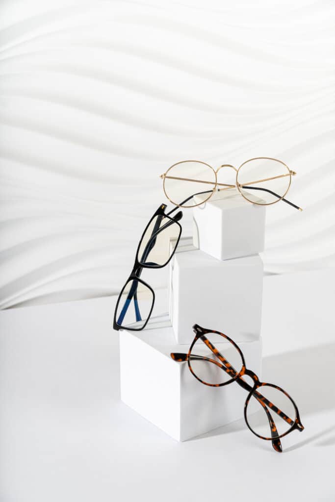 Trois paires de lunettes de vue (dorées rondes, noires rectangulaires, écaille rondes) sur des podiums blancs épurés.