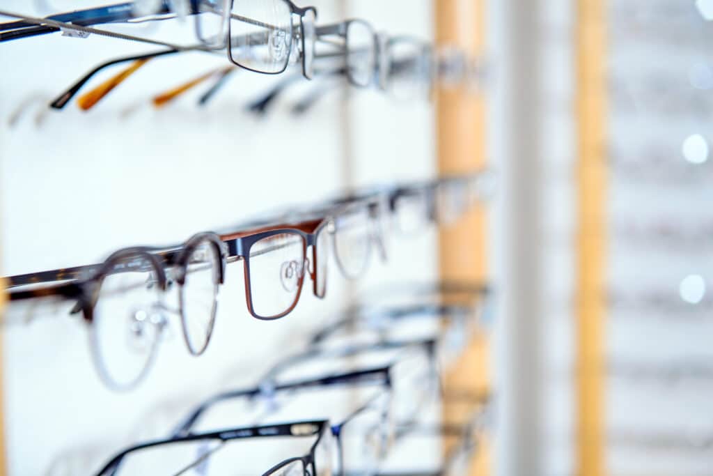 Plusieurs rangées de lunettes de vue sur un présentoir mural, focalisant sur une paire à monture foncée avec reflets.