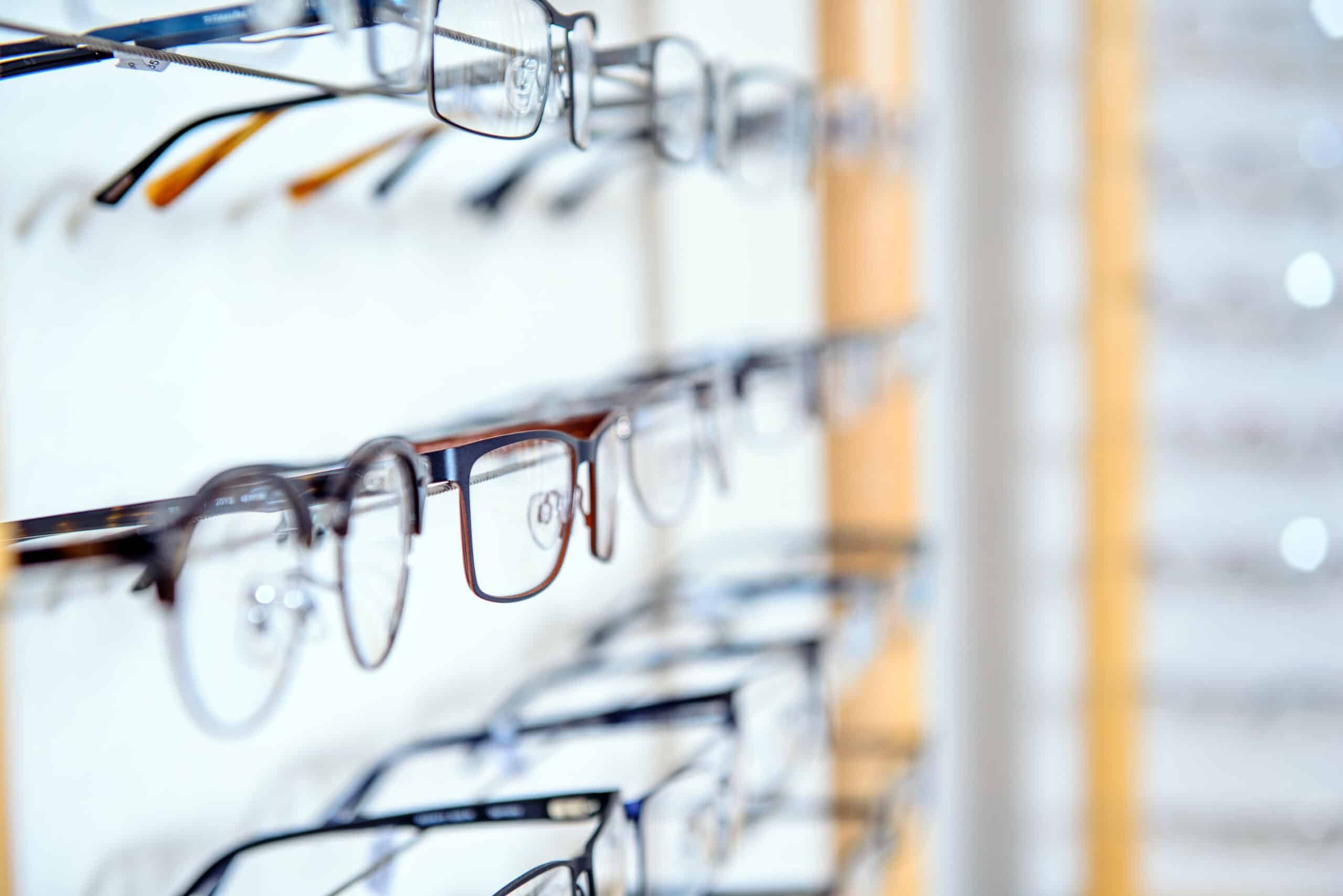 Plusieurs rangées de lunettes de vue sur un présentoir mural, focalisant sur une paire à monture foncée avec reflets.