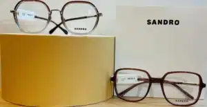 Deux paires de lunettes SANDRO. Une monture ronde métallique à 198€, et une monture carrée marron à 195€. Logo SANDRO visible sur les lentilles et une boîte blanche.