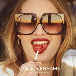 Femme souriante en lunettes de soleil tendance Daniel Benhams, suçant un lollipop rouge. Reflets de coucher de soleil. GLAMOUROUS EYEWEAR.