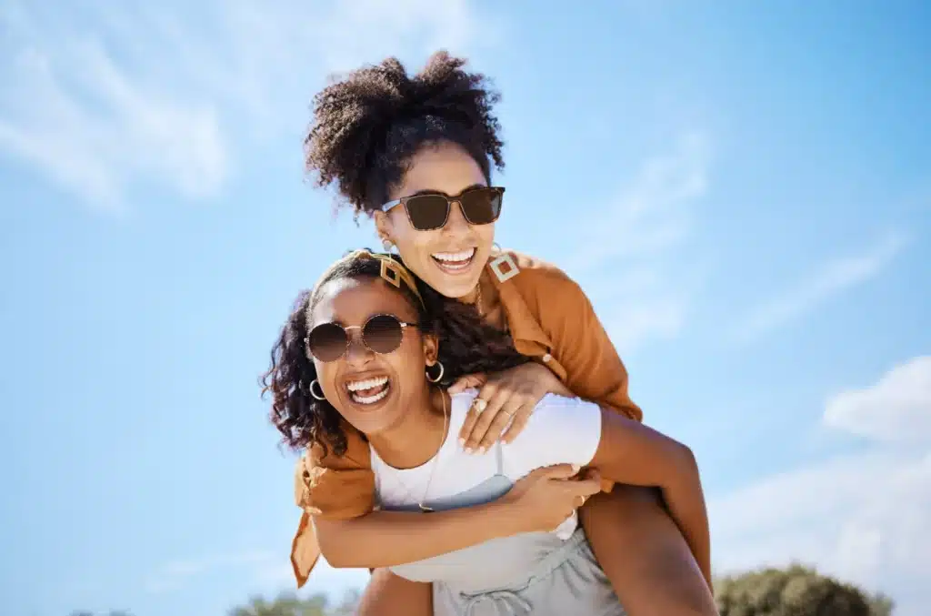 Deux amies rient, l'une sur le dos de l'autre, toutes deux avec des lunettes de soleil, sous un ciel bleu d'été. Scène de joie en plein air.