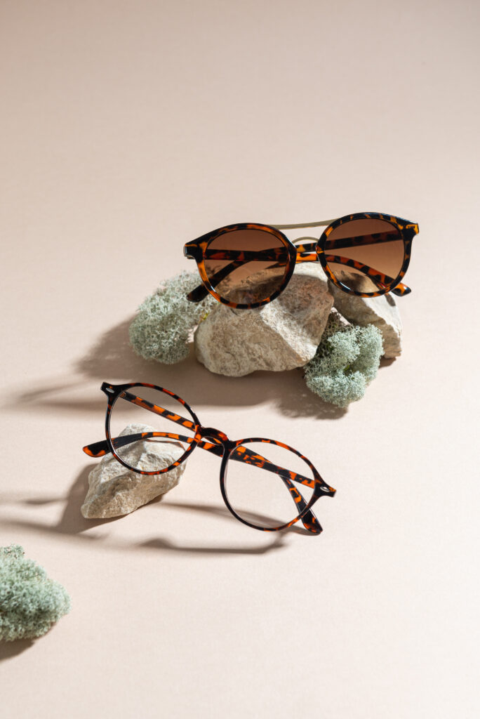 Lunettes de soleil et lunettes de vue écaille, posées sur roches et mousse naturelle sur fond beige. Lumière naturelle et ombres.